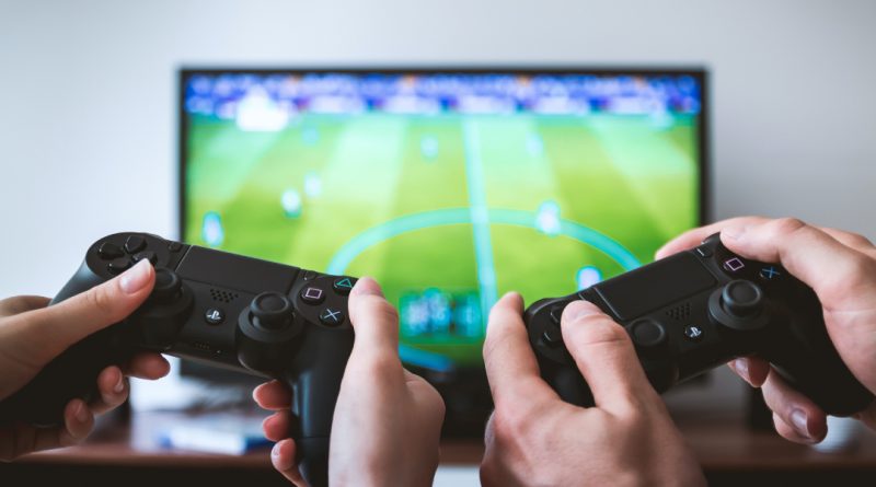 Two PS4 controllers in teh foregrund, held up in front of a TV showing FIFA